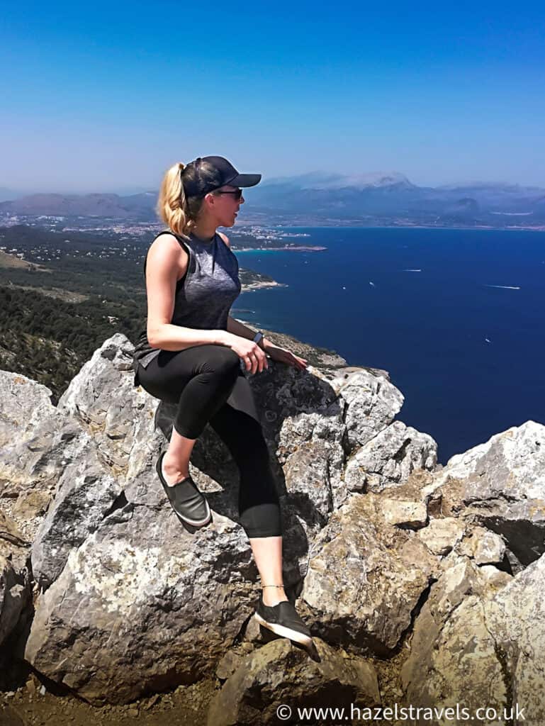 Woman with blonde hair sits on the top of a mountain in Mallorca, Spain, overlooking the Mediterranean sea.