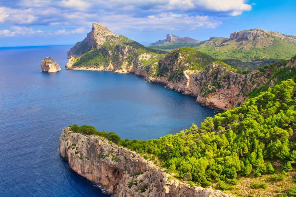 A rocky coastline with steep cliffs and dense green forest meets a calm blue sea under a partly cloudy sky.