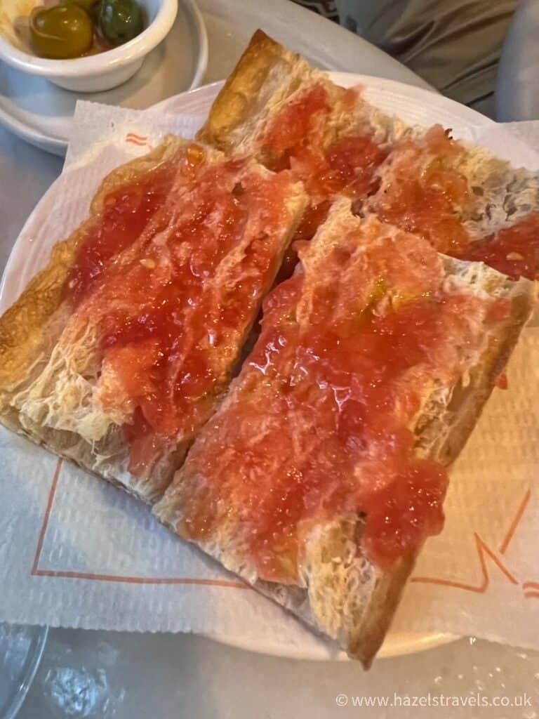 A plate features Pain con tomate, where slices of bread are artfully topped with crushed tomato, while a small bowl of olives sits invitingly in the background.