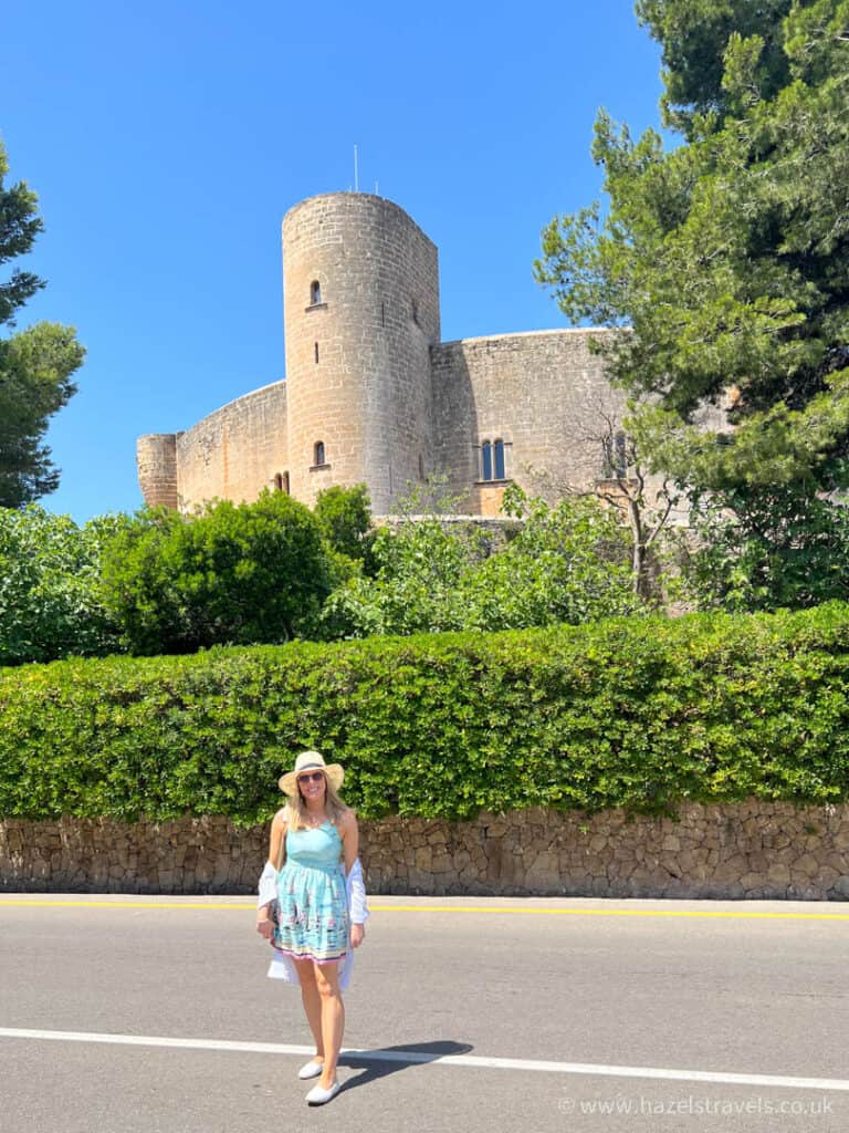 A woman in a light blue dress and hat stands on a road in front of a stone castle surrounded by greenery on a sunny day.