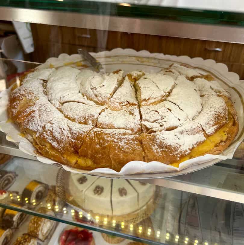 A round, sliced pastry dusted with powdered sugar is displayed on a white paper doily inside a glass bakery case.