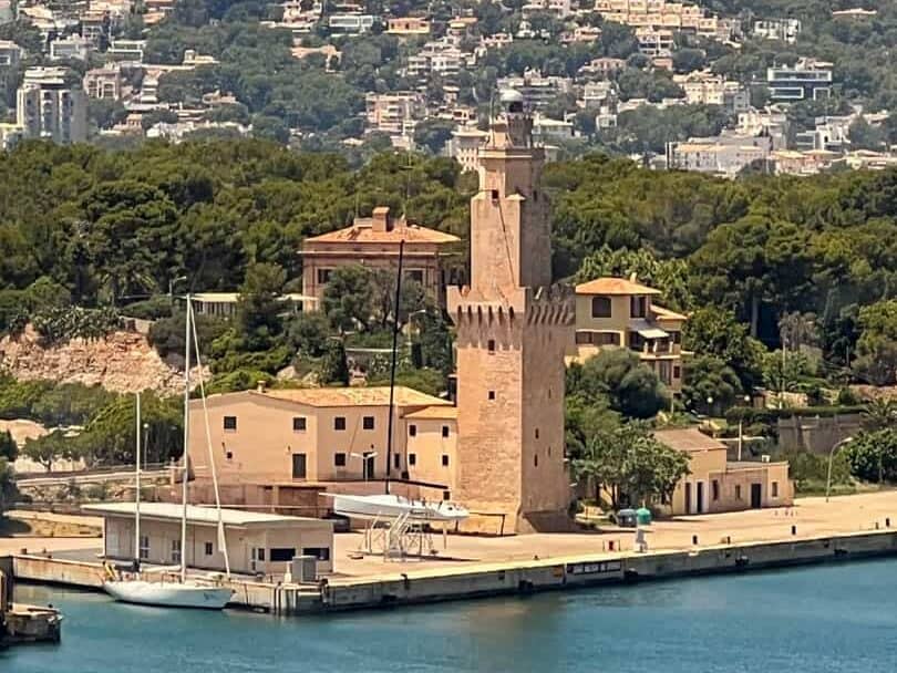 A stone lighthouse with attached buildings stands near a waterfront, surrounded by trees, with a town visible in the background.