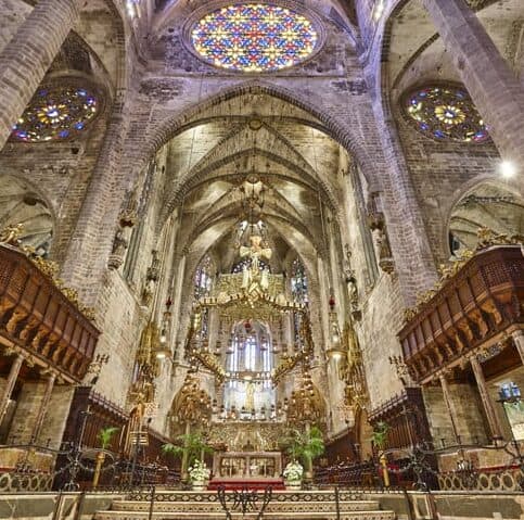 Interior view of a grand Gothic cathedral with high vaulted ceilings, stained glass rose windows, ornate chandeliers, and elaborately decorated altar area.