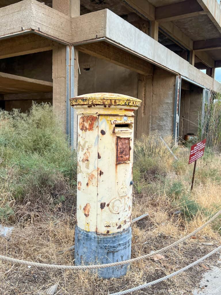 A weathered, rusted post box stands in front of an abandoned building, surrounded by overgrown grass and weeds