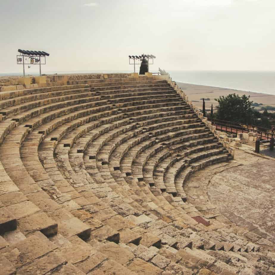 Stone amphitheater with curved, tiered seating overlooking a distant coastline under a hazy sky.