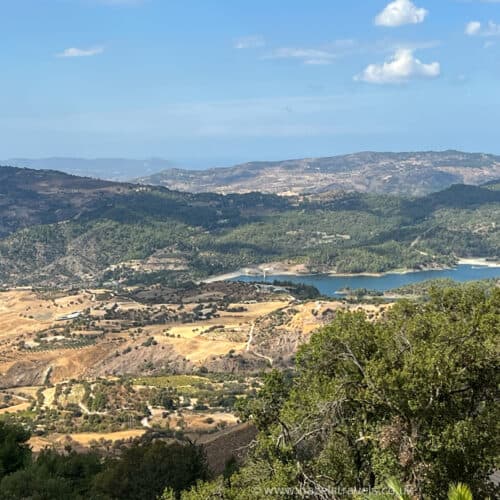 A scenic landscape of the Troodos Mountains in Cyprus featuring rolling hills, patches of green forest, a small lake, and a partly cloudy sky