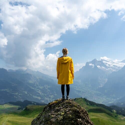 Woman in a yellow coat stands on top of a rock and looks out over mountain peaks and clouds