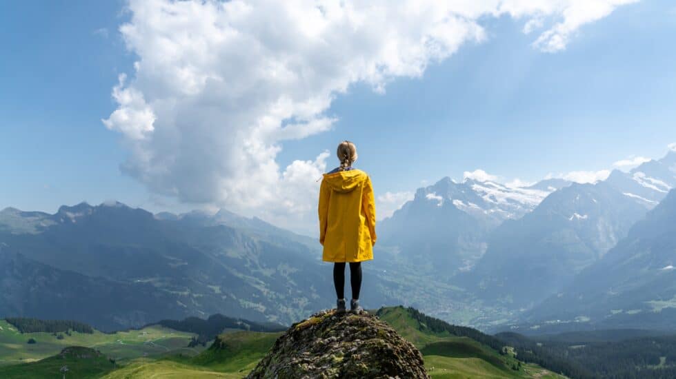 Woman in a yellow coat stands on top of a rock and looks out over mountain peaks and clouds