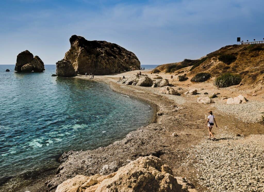 A person strolls along a rocky beach in Cyprus, with majestic rock formations rising from the sea and a clear sky stretching overhead.