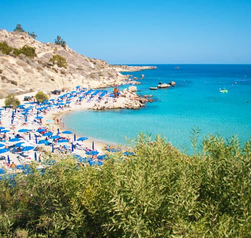 Sandy beach with blue umbrellas and loungers along a turquoise shoreline, surrounded by rocky hills and greenery under a clear blue sky—a perfect scene typical of the breathtaking beaches in Cyprus.