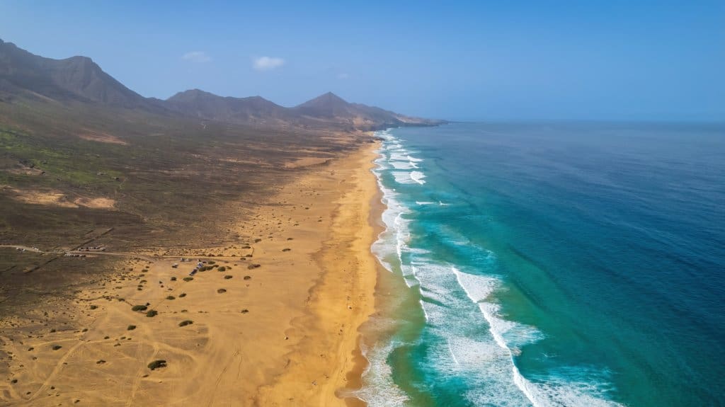 Aerial view of a long sandy beach with waves meeting the shore, bordered by mountains and the ocean beneath a clear sky—discover this stunning scene in our Fuerteventura Travel Guide.