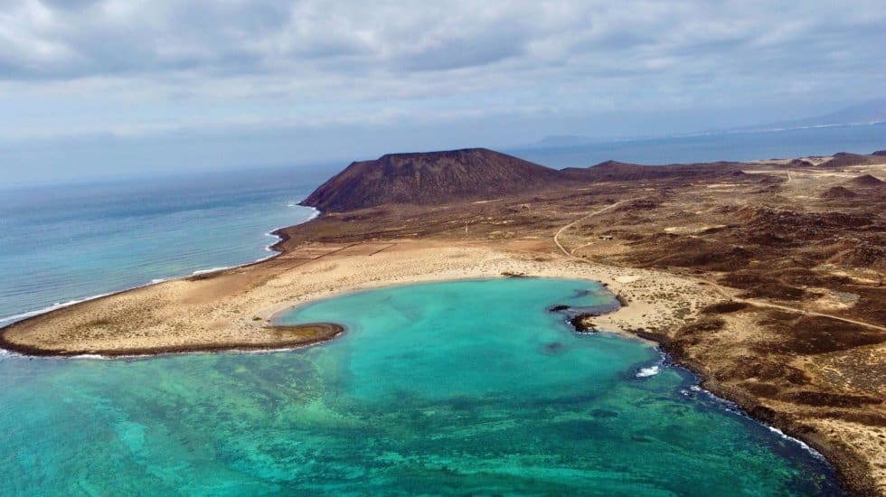 Aerial view of Lobos Island with blue/green water, crescent shaped yellow sand beach and large brown volcano caldera in the distance.