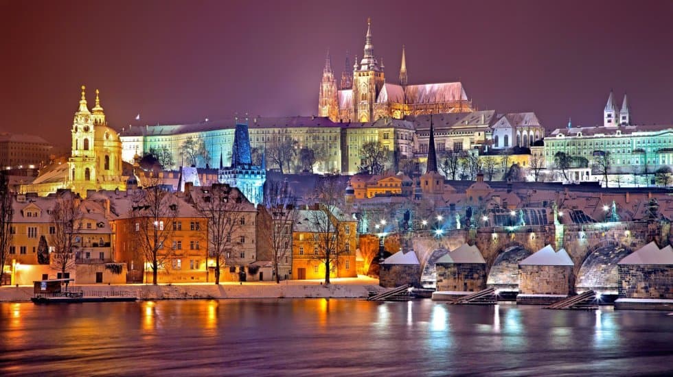 Pretty picture postcard scene depicting a European city (Prague) lit up in various pastel colours. View from over a river, with a castle with spires on top of a hill in the distance.