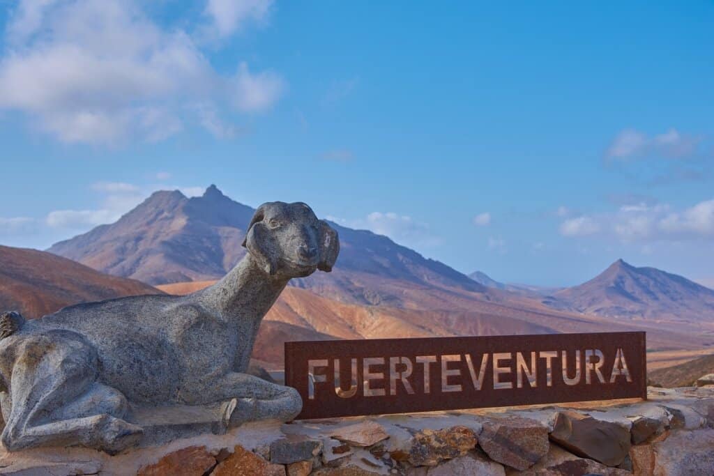 Fuerteventura Welcome Sign with a Canarian goat sculpture. Blue sky and brown/red mountainous landscape in the background.