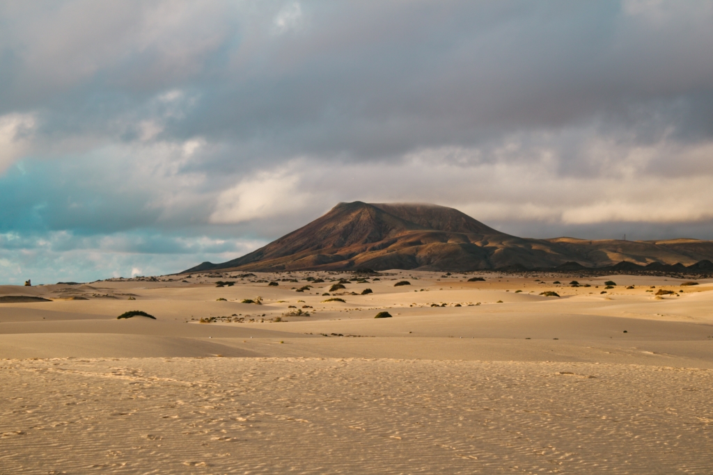 Sandscape with yellow sand and small dues, cloudy sky with brown volcanic mountain on the horizon.