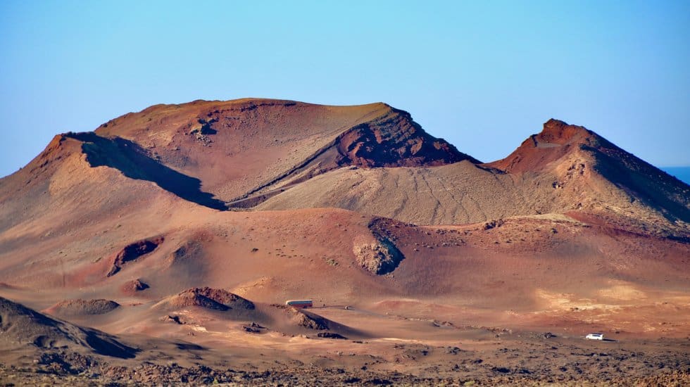 Huge red and brown volcanic mountains rising up into a bright blue sky. Two tiny buses can be seen in the foreground driving along the road to the crater of Mount Timanfaya in Lanzarote.