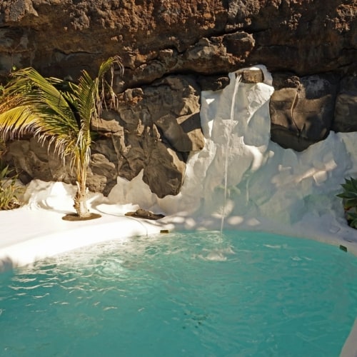A small green pool surrounded by white painted rocks and brown volcanic rock, green foliage and palms, small white fountain pouring into the pool.