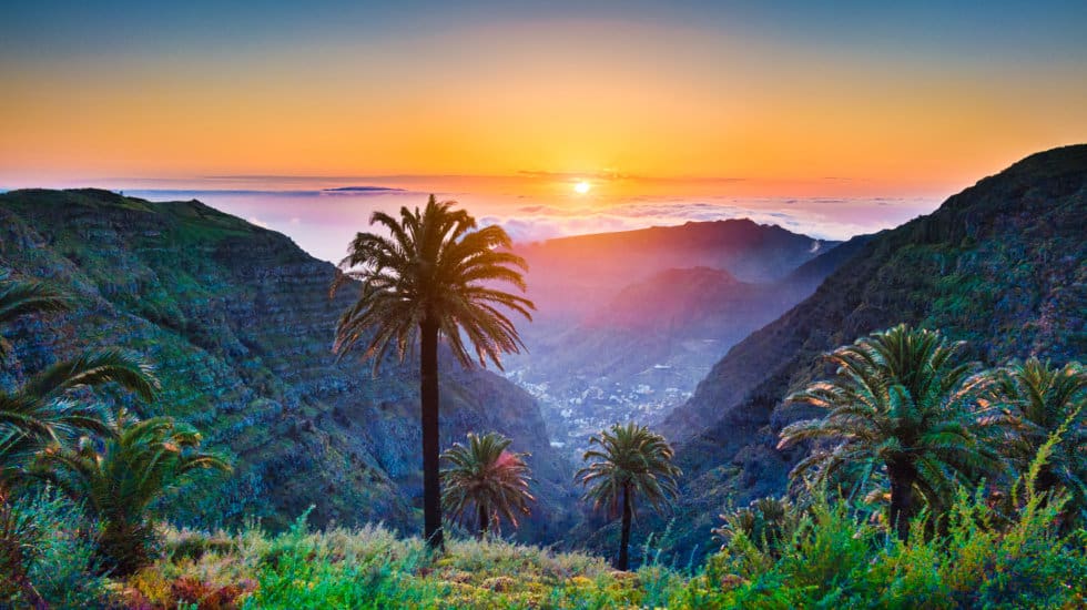 Which Canary Island Is the Best: Sunset view over lush green mountains and palm trees, with white clouds in the disitance and a setting sun.