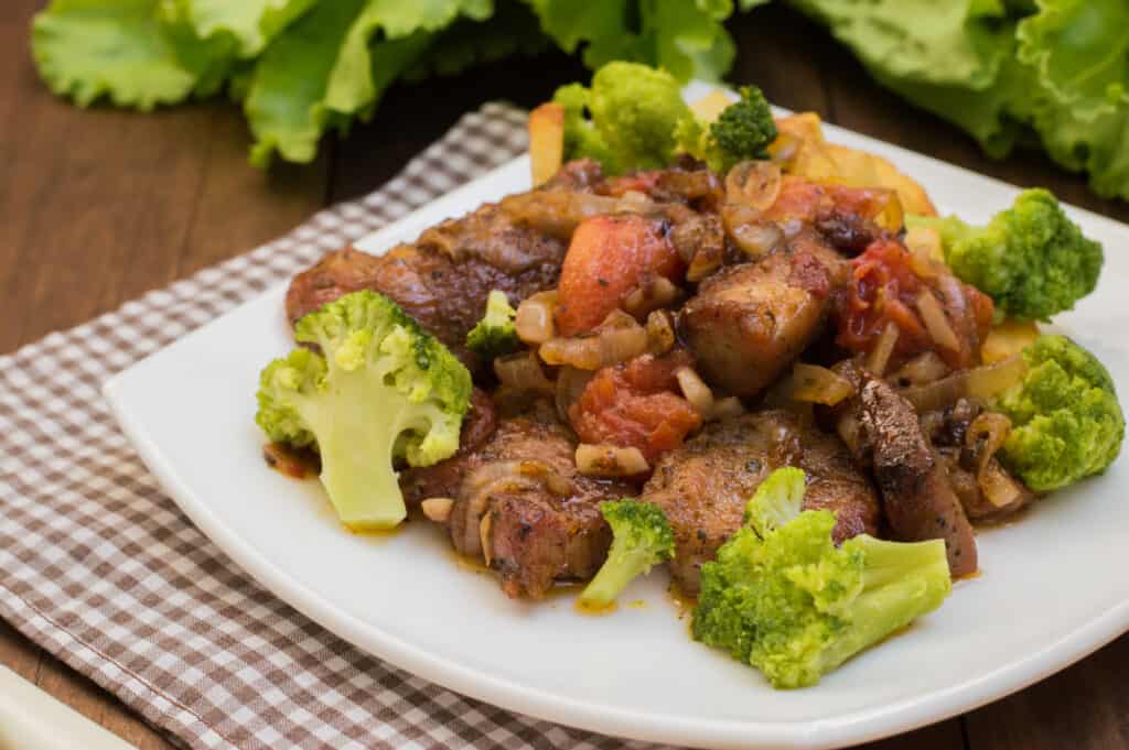 A plate of stir-fried beef and broccoli, reminiscent of a modern stifado, mingles with tomatoes and onions on a checkered cloth, while leafy greens in the background add a fresh flourish.