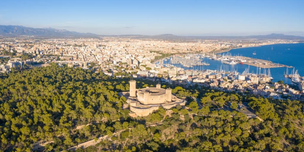 Aerial view of Bellver Castle nestled among trees, with the cityscape and Palma harbour nearby under a clear blue sky.