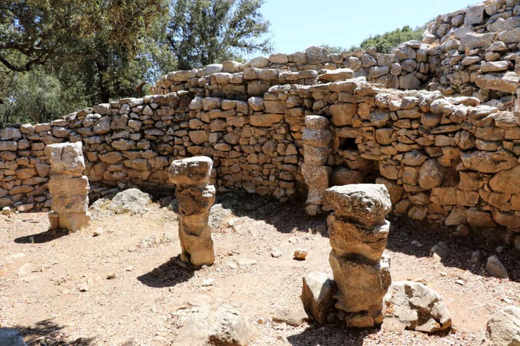 The ancient stone ruins of Poblat Talaiotic de Ses Paisses near Arta feature three pillar-like structures in the foreground, with a stone wall and doorway in the background, surrounded by scattered rocks and trees.