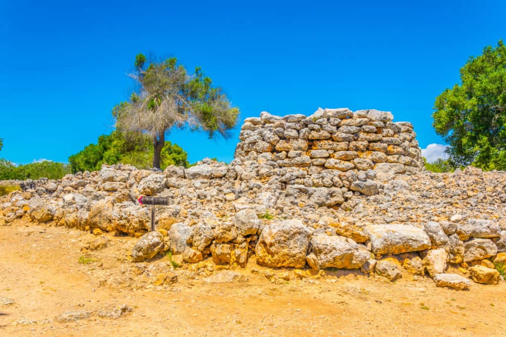 Under a clear blue sky, the ancient stone structure of the Ruins of Talayot Capocorb Vell rises majestically, surrounded by trees and dry ground. A small signpost stands nearby, silently narrating tales of history long past.