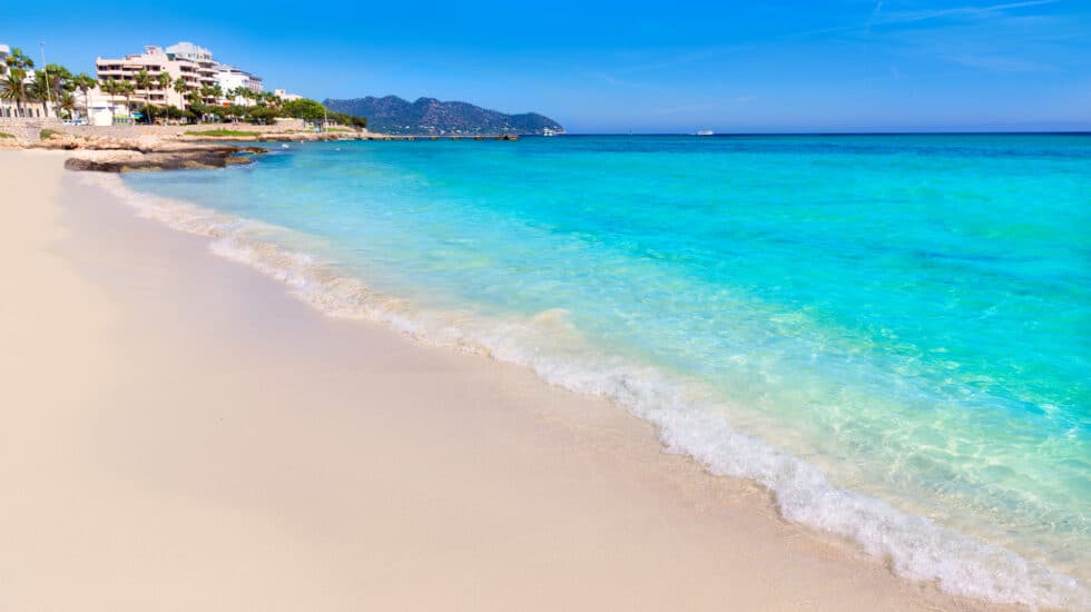 The sandy beach of Cala Millor boasts clear turquoise waters under a bright blue sky, with charming buildings and hills providing a picturesque backdrop.