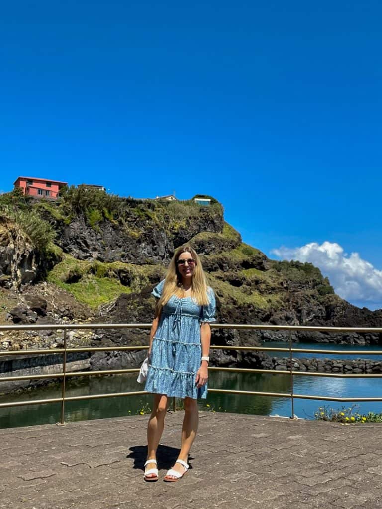 Woman in a blue dress standing in front of a cliff with a house on top under a clear blue sky.