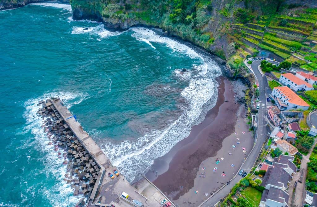 Aerial view of a curved beach with dark sand beside a cliff, with a breakwater and buildings nearby. Part of an itinerary for Madeira.
