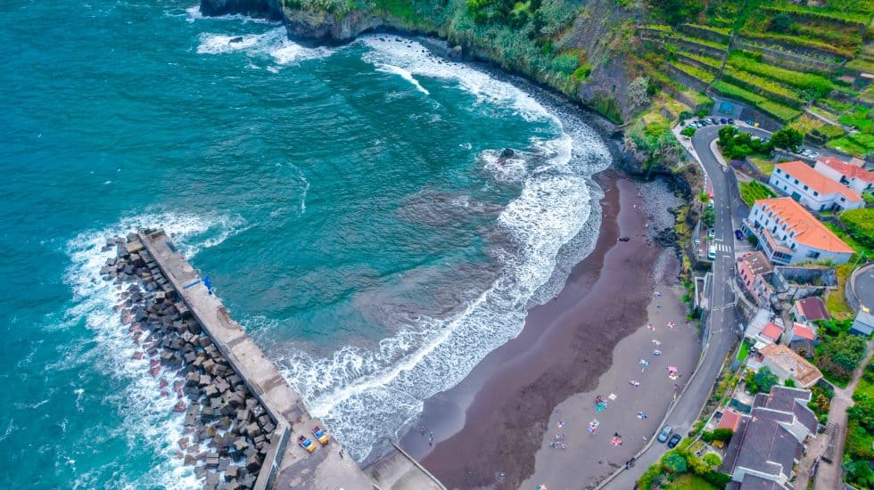 Aerial view of a curved beach with dark sand beside a cliff, with a breakwater and buildings nearby. Part of an itinerary for Madeira.