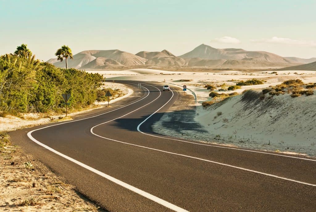 A winding paved road runs through a desert landscape with sparse vegetation and distant mountains under a clear sky. Two cars are seen on the road.