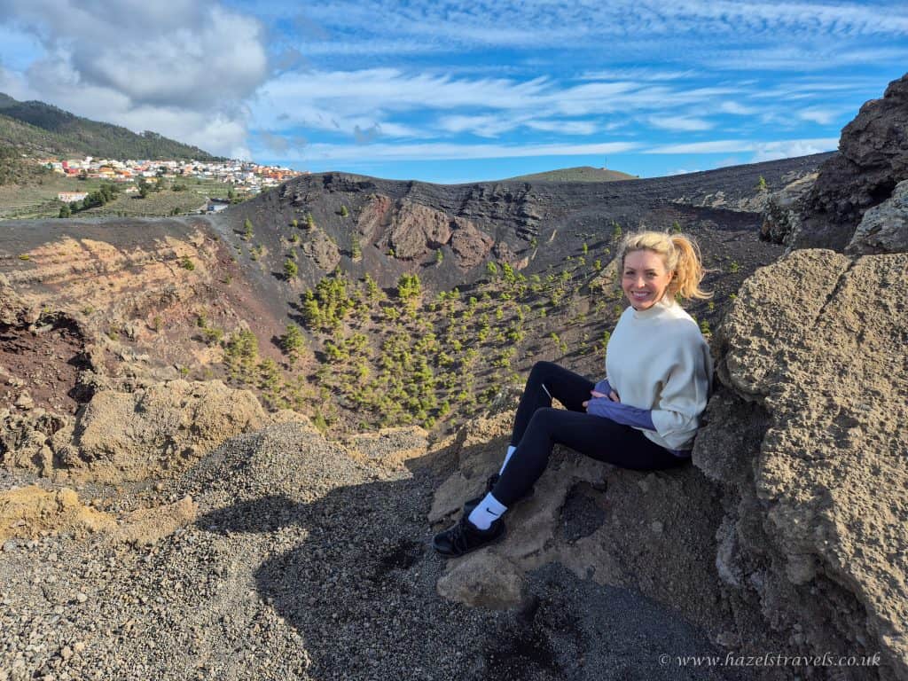 A woman sits on a rocky ledge overlooking a volcanic landscape with scattered trees and a distant village under a partly cloudy sky.