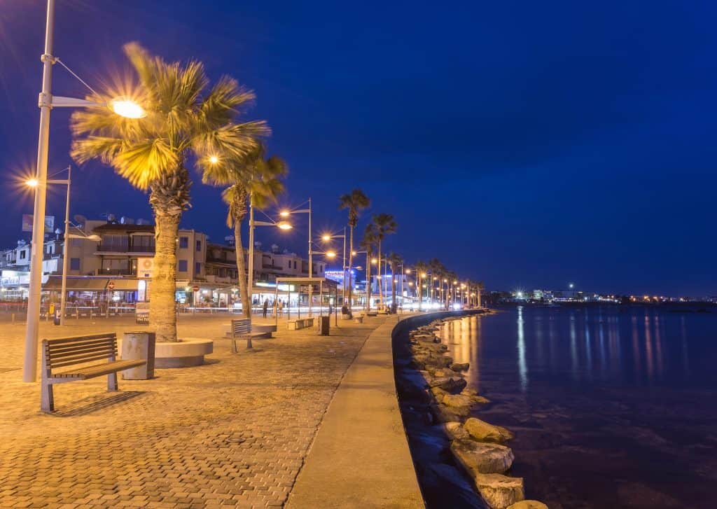 Palm-lined promenade in Paphos, Cyprus, with benches and streetlights at night, next to calm water and buildings with lit storefronts in the background.