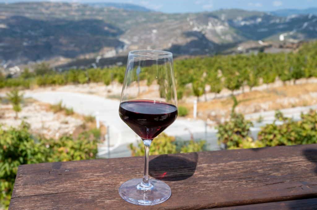 A glass of red wine on a wooden table with a vineyard and the Troodos mountains in Cyprus in the background under a clear sky.