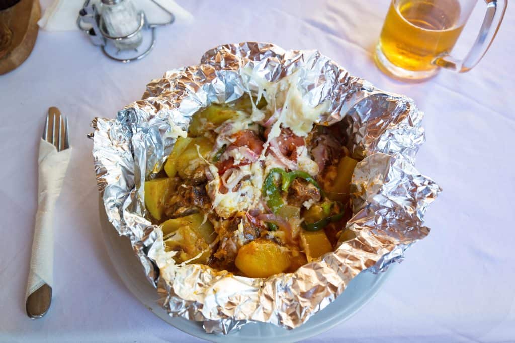 A Cypriot baked dish of meat (kleftiko) potatoes, vegetables, and cheese served in aluminum foil on a plate, with a fork, napkin, and glass of beer on a white tablecloth.