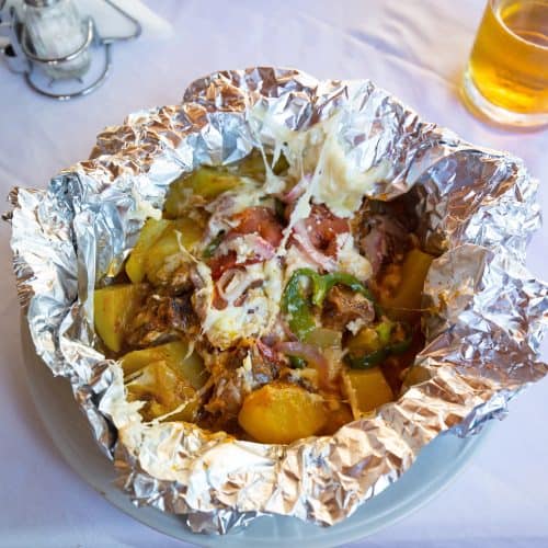 A Cypriot baked dish of meat (kleftiko) potatoes, vegetables, and cheese served in aluminum foil on a plate, with a fork, napkin, and glass of beer on a white tablecloth.