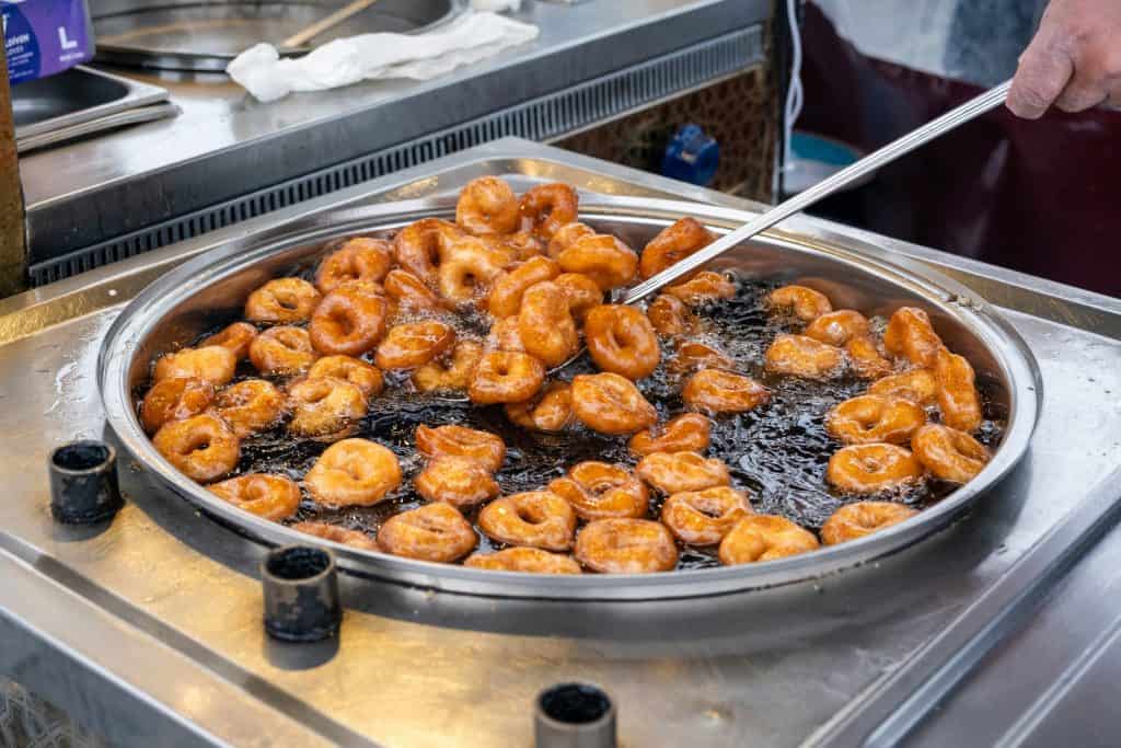 A large metal pan filled with small ring-shaped doughnuts frying in oil, with a person stirring them using a metal tool.