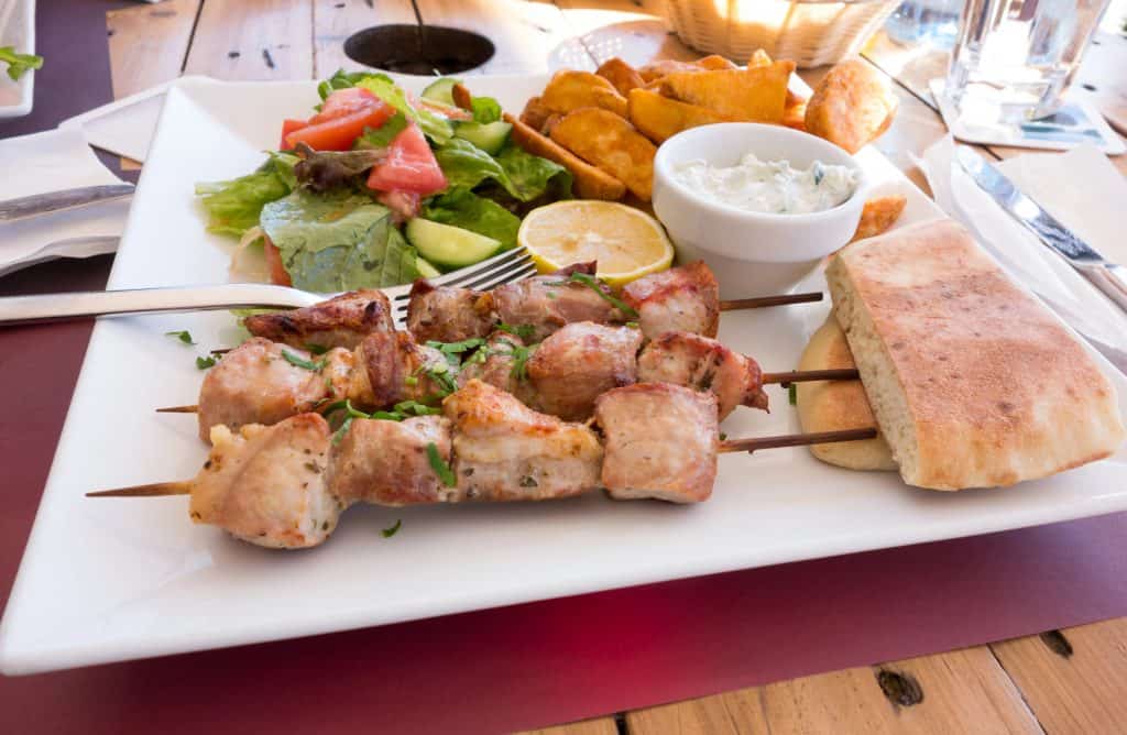 A plate with grilled meat skewers, salad, potato wedges, pita bread, a lemon wedge, and a small bowl of dip on a wooden table.