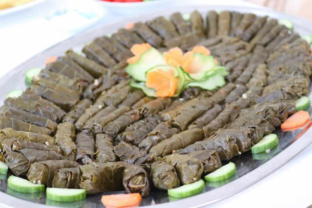 A platter of stuffed vine leaves arranged in a circular pattern, garnished with cucumber and carrot slices, with carved vegetables in the center.