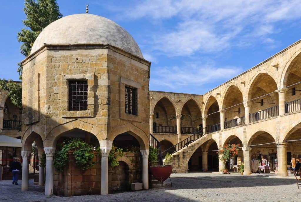 A historic stone courtyard with arched walkways and a domed structure at the center, surrounded by columns and greenery under a partly cloudy sky. The famous Buyak Han in Nicosia, Cyprus.