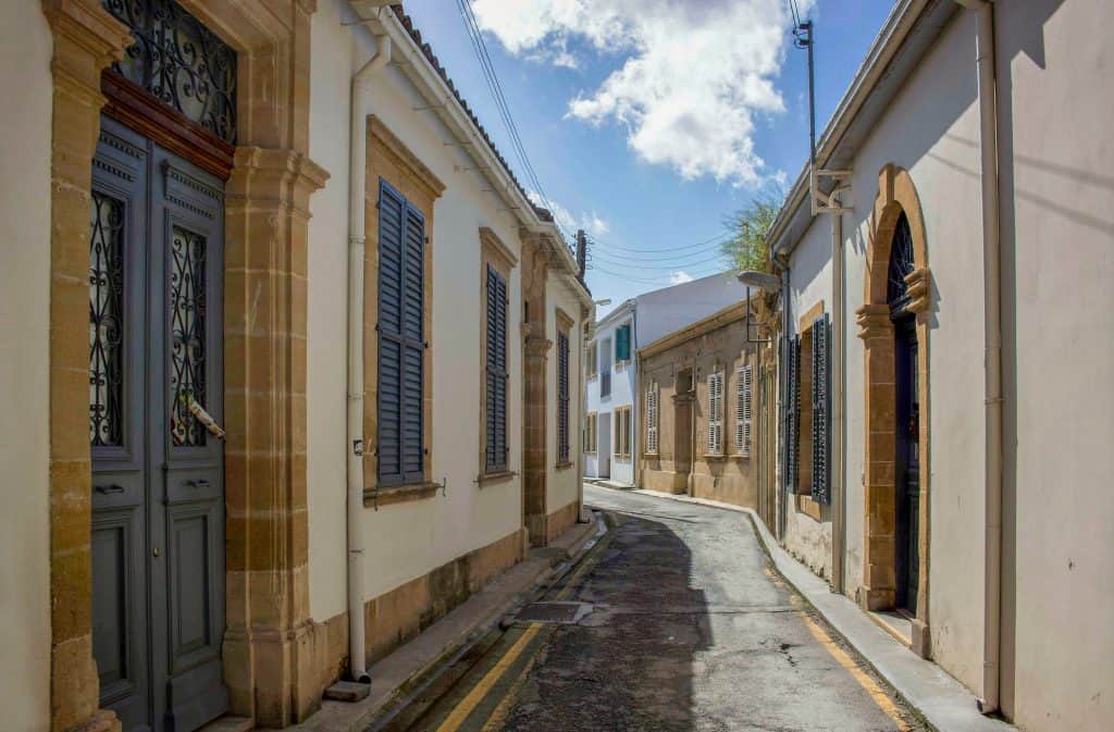 Narrow street in Nicosia Cyprus lined with traditional Mediterranean-style buildings, featuring shuttered windows and stone arches, under a partly cloudy sky.