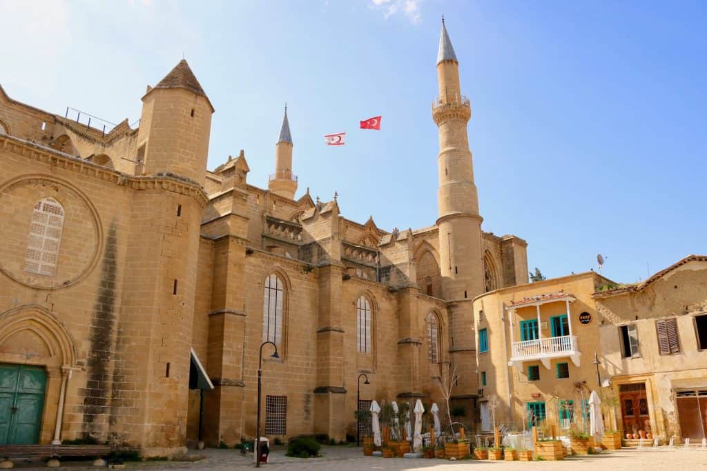 A large historic stone mosque in Nicosia, Cyprus, with minarets and arched windows, two flags flying above, and a small cafe with outdoor seating in the foreground.