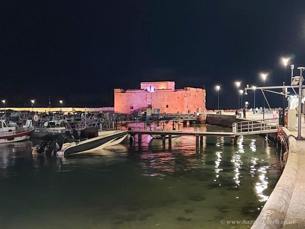Night view of a waterfront fort in Paphos, Cyprus illuminated with purple lights, surrounded by small boats and piers, with streetlights reflecting on the water.