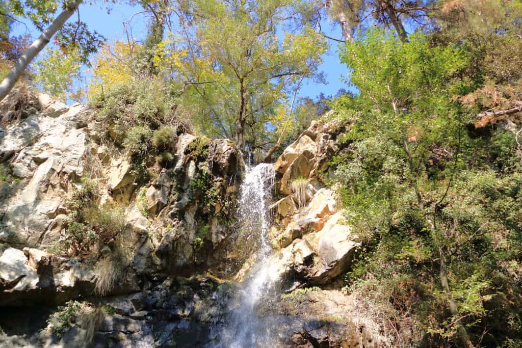 A small waterfall cascades down a rocky cliff in the Troodos mountains in Cyprus, surrounded by green trees and foliage under a clear blue sky.