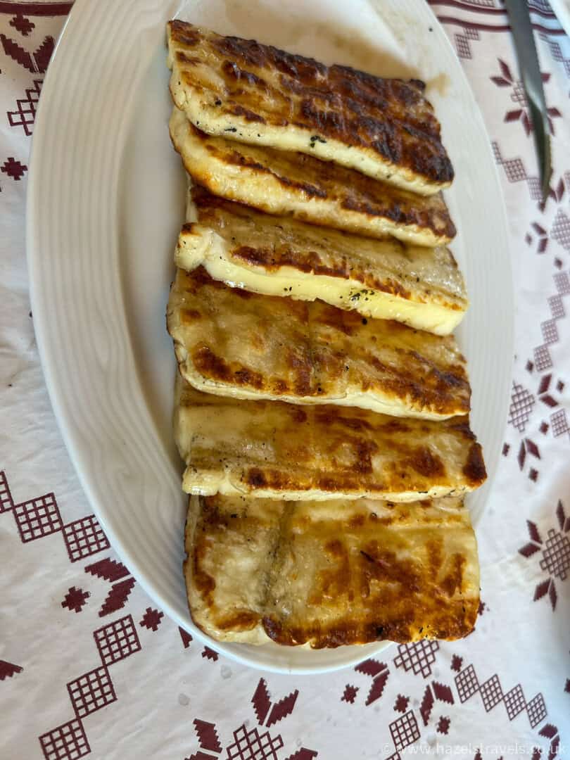 Six rectangular pieces of grilled halloumi cheese arranged in a row on a white oval plate, placed on a patterned tablecloth.