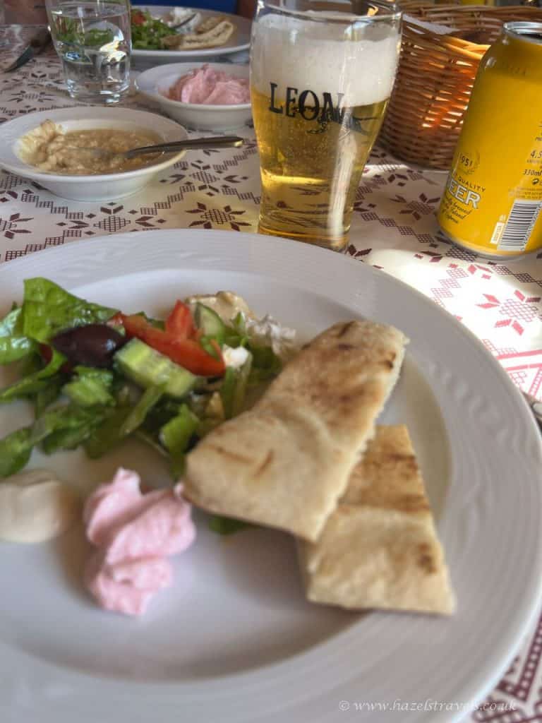 A plate with pita bread, salad, and dips sits on a table next to a glass of beer, a can, and additional plates of food in the background.