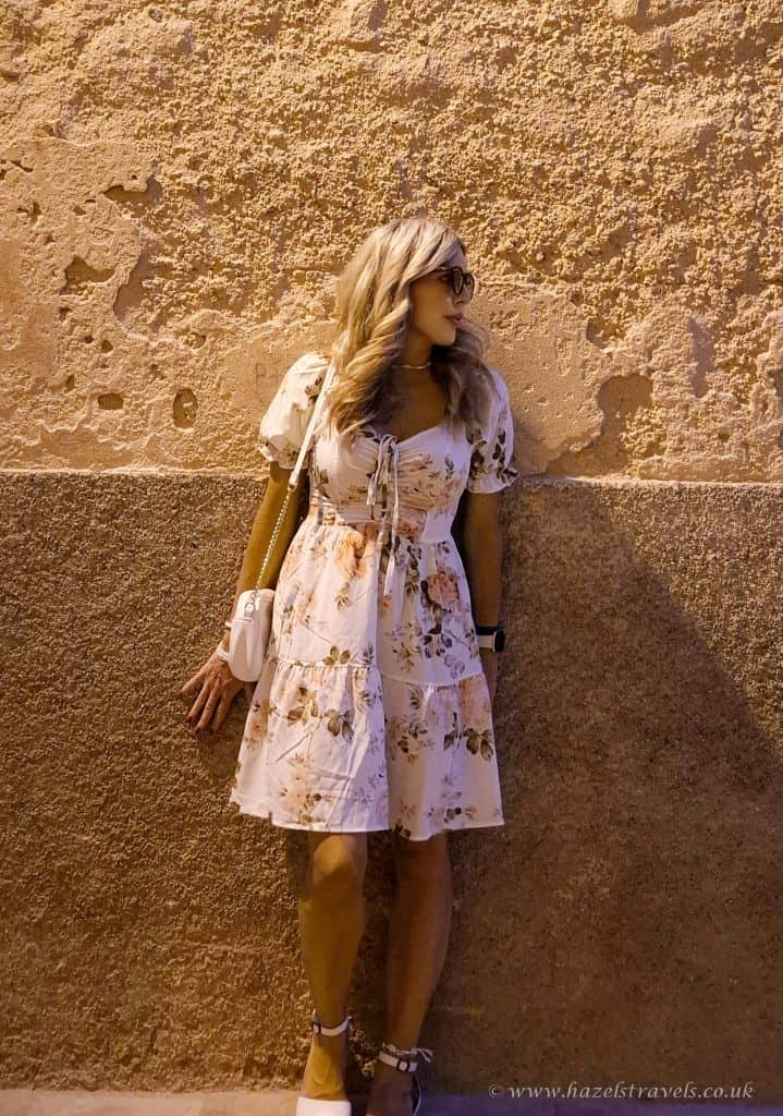 Stylish woman in floral summer dress with sunglasses standing against textured terracotta wall - perfect travel fashion for sunny destinations.