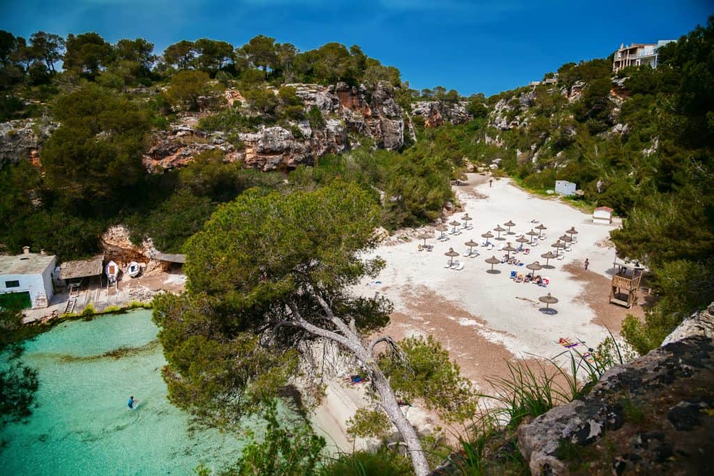 A small sandy beach with clear turquoise water, surrounded by rocky cliffs and trees, with sun loungers and umbrellas scattered along the shore.