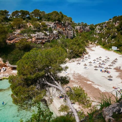 Turquoise beach with sun umbrellas in a secluded cove, surrounded by rocky cliffs and lush greenery, perfect for relaxing holidays and coastal exploration in the Mediterranean.