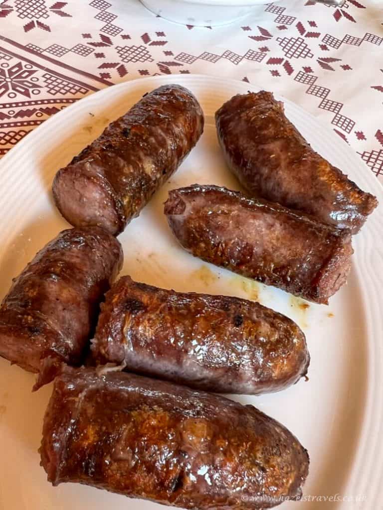 Six grilled sausages are arranged on a white plate, placed on a patterned tablecloth.